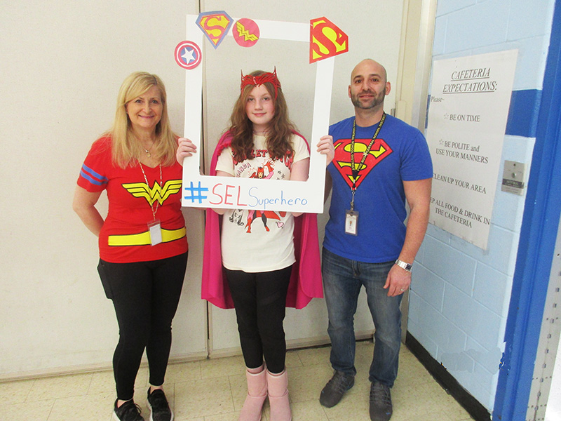 Two adults stand on either side with of a middle school girl. they all have super hero outfits on and the person in the middle holds up a frame with super hero logos on it.