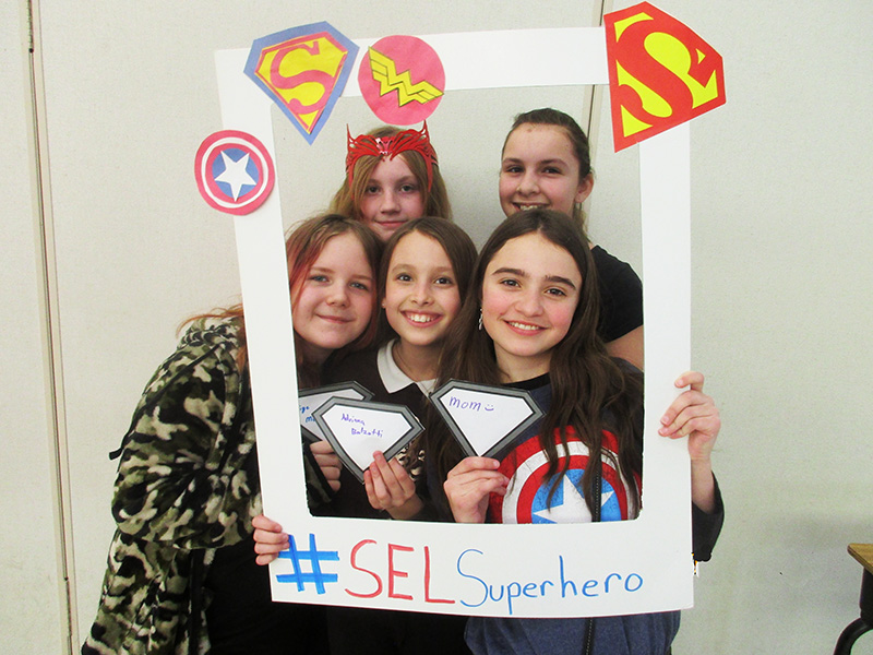 Five middle school girls smile as they hold up a photo frame with all superhero logos on it.
