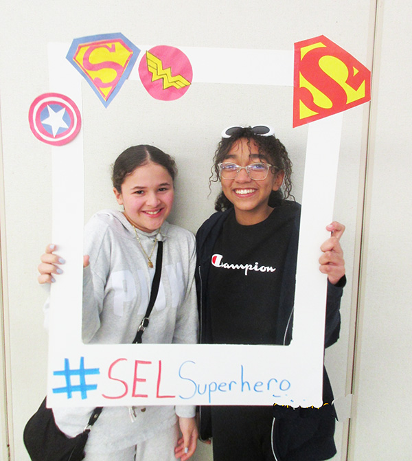 Two middle school students smile as they hold up a frame with all super hero symbols on it.