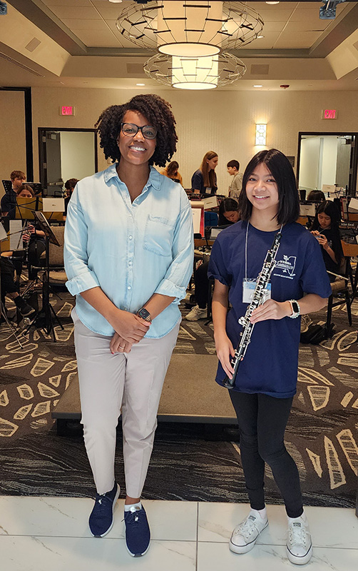 A young middle school woman with long dark hair, wearing a blue shirt and black pants holding an oboe, stands with a woman with chin-length dark hair. Both are smiling.