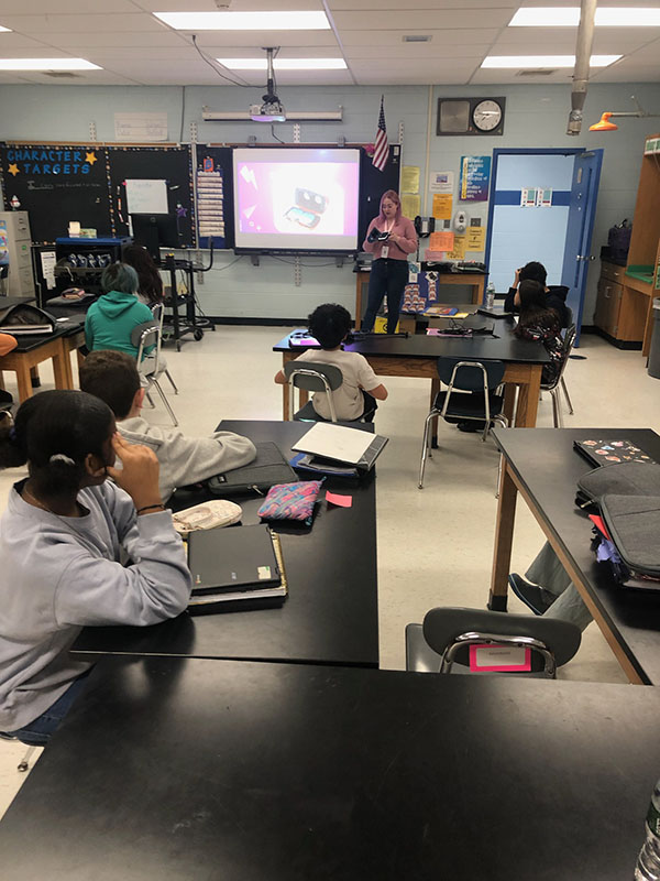 A classroom setting with middle school student sitting at tables looking toward the front of the room. A woman is in front, pointing to a screen.