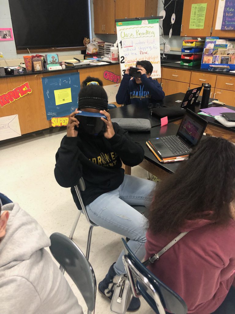 Middle school students are sitting at a table looking through virtual reality goggles.