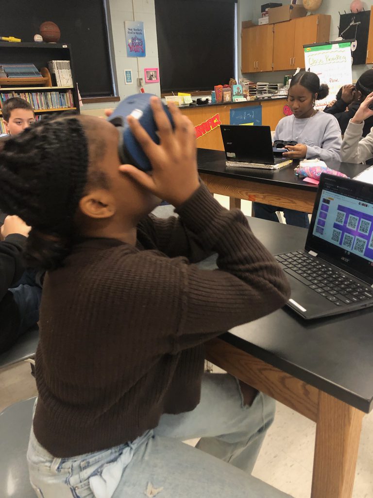 A middle school student sits at a table and looks through virtual reality goggles.