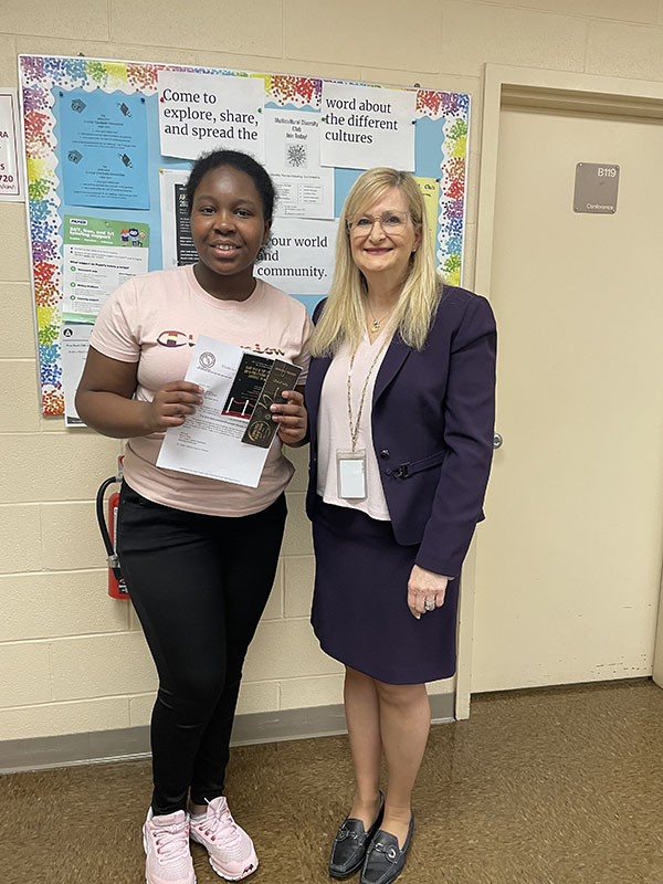 A young woman in black pants and peach colored shirt smiles and holds a booklet in front of her. Her dark hair is pulled back. She is standing with a woman wearing a blue suit and white blouse, long blonde hair. She is smiling too.