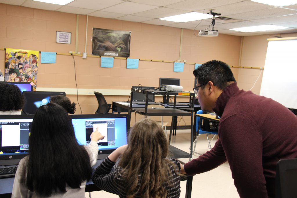 A classroom setting with tables filled with computer screens. A man with short dark hair, glasses, and wearing a maroon sweater leans over to help two students sitting at the computers.