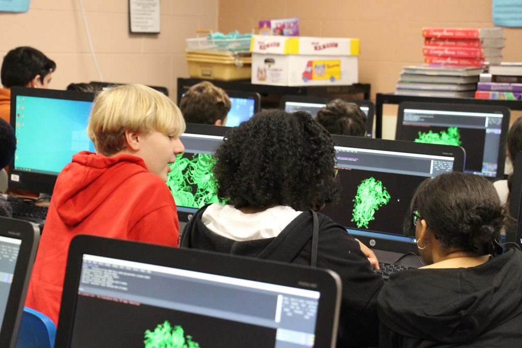 Students sit looking at a computer screen that shows a green structure being built.