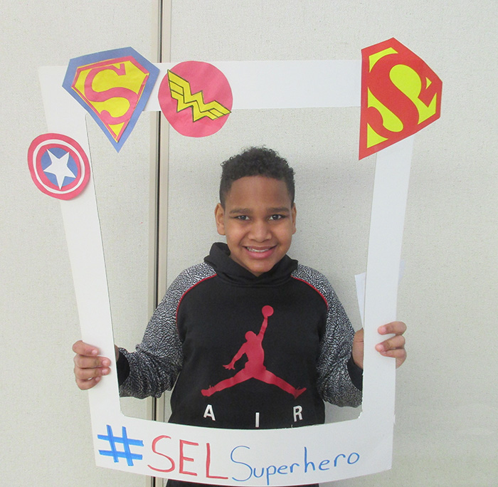 A middle school student smiles as they hold up a frame with all super hero symbols on it.