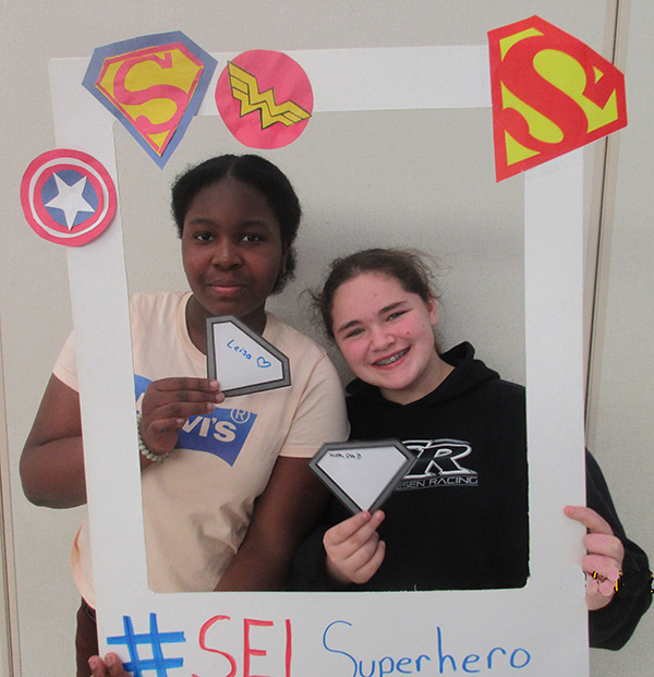 Two middle school students smile as they hold up a frame with all super hero symbols on it.