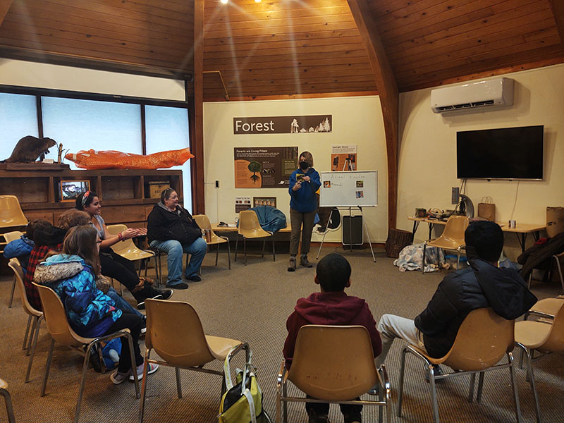 A circle of chairs with middle school kids sitting watching an instructor talk to them.