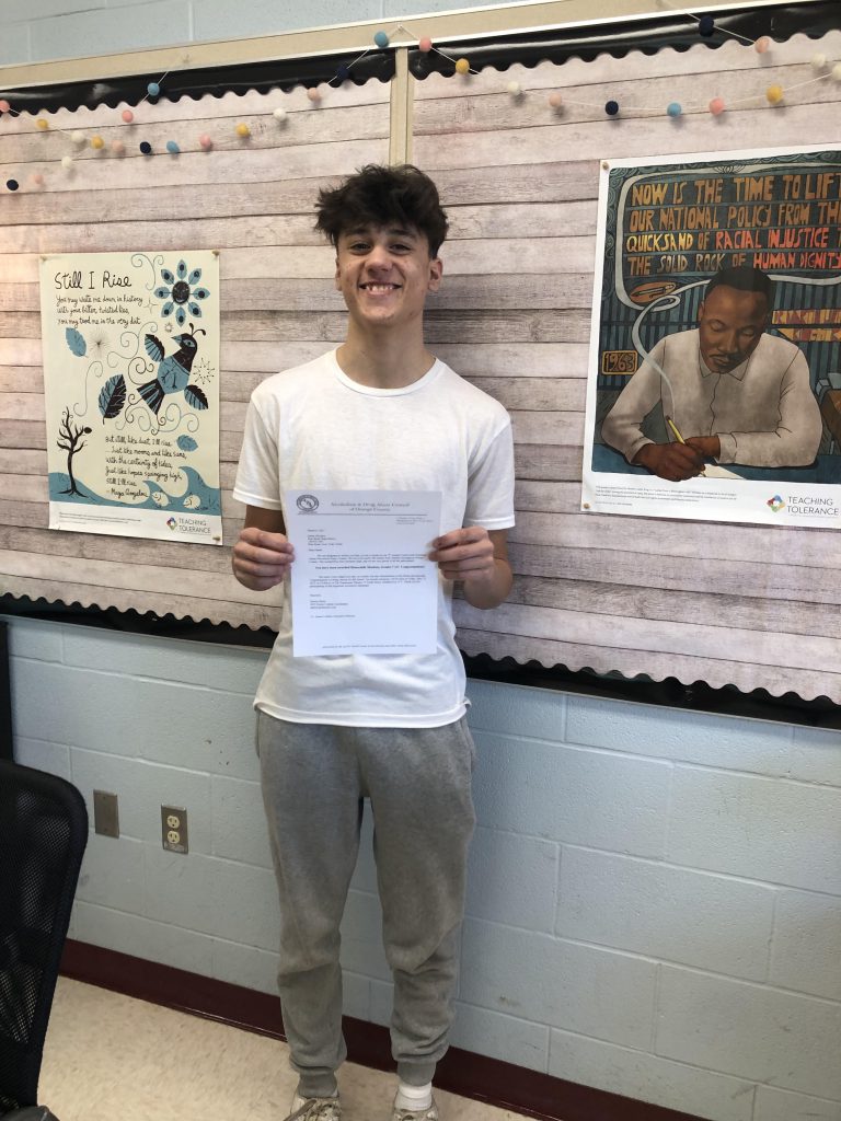 A high school young man with dark hair,white shirt and tan pants stands smiling proudly.