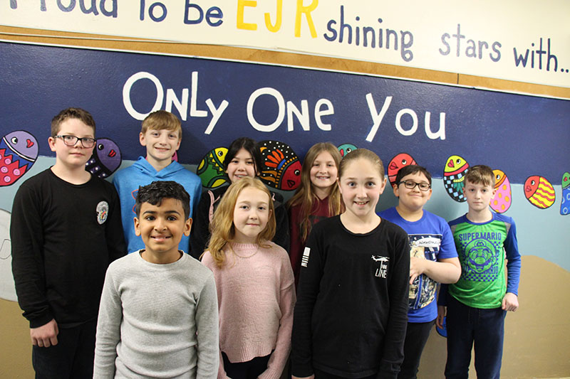 A group of nine older elementary students stand together smiling. The background says Proud to be EJR shining star. There's only one you.