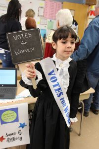 A third-grade girl with dark hair is wearing an old-fashioned black and white long dress. She has a sash that says Votes for Women and is holding a sign that says Votes for women. She is Susan B. Anthony.