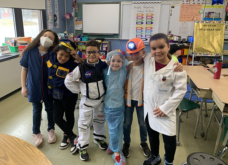 A group of five second grade students stand arm-in-arm, dressed in clothes of different professions - there is a pharmacist, construction worker, dentist, astronaut and police officer. There is an adult on the left.