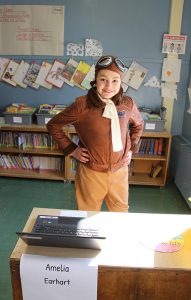 A third-grade girl dressed in a brown leather jacket and scarf with goggles sitting on top of her head.  She is Amelia Earhart.