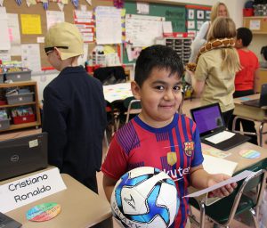 A third-grade boy holds a soccer ball and is wearing a red and blue soccer jersey. He is Cristiano Ronaldo.