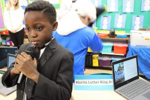 A third-grade boy is dressed in a dark suit and white shirt. He is holding a microphone. He is Martin Luther King.