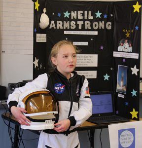 A third grade girl is dressed in a white astronaut uniform, holding a helmet. She is Neil Armstrong.