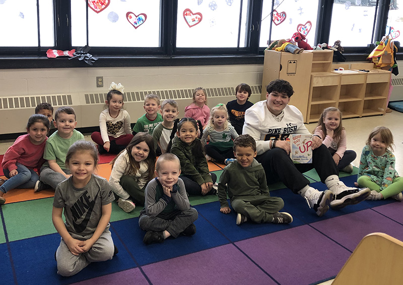 A high school student wearing a white football jersey sits with a group of young elementary kids after reading to them.