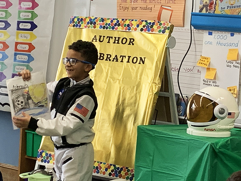 A second grade boy is dressed in a white astronaut suit as he shows a book to an audience in a classroom.