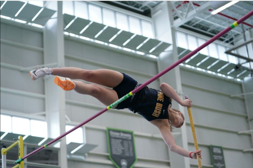 A high school pole vaulter in a navy blue uniform with Pine Bush written on it goes over the pole.