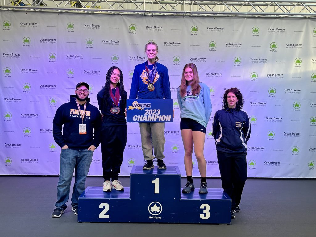 Three high school young women track athletes stand on a podium with steps numbered 1, 2 and 3. On either side is an adult, a man on the left and a woman on the right.