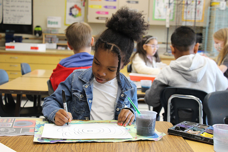 A second-grade girl wearing a white shirt, denim jacket with her hair pulled up into a high bun, draws on paper.