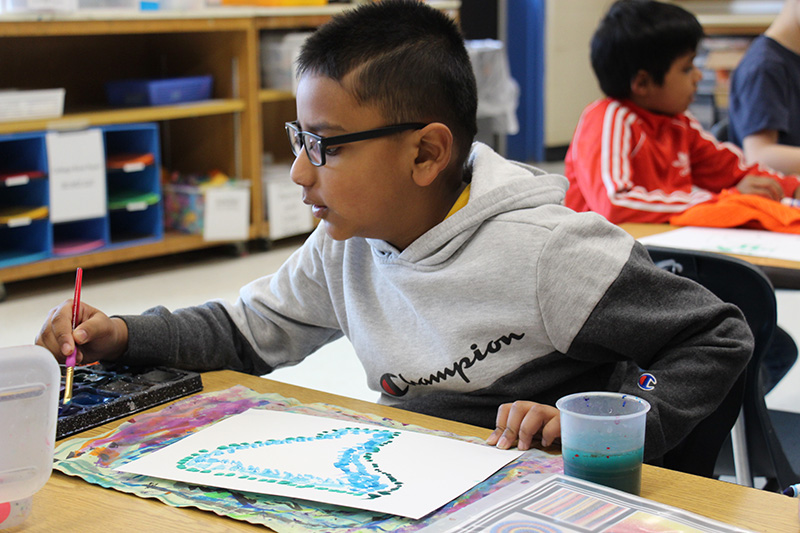 A second-grade boy with short dark hair, glasses and wearing a gray sweatshirt leans over to dip his brush into paint.