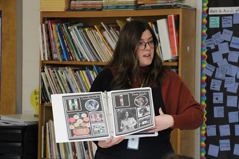 A woman with long dark hair and glasses, wearing a maroon sweater, holds up a binder of different artwork.