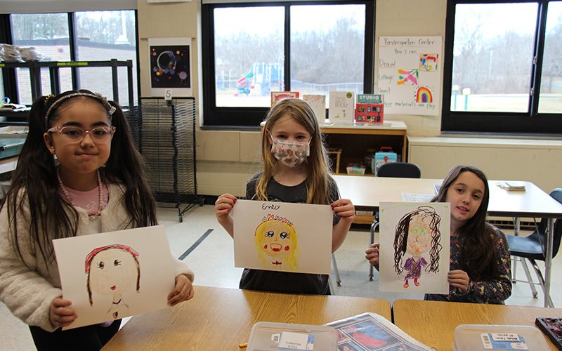 Three second-grade girls hold up their self portraits they painted.