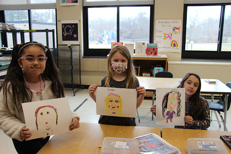 Three second-grade girls hold up their self portraits they painted.