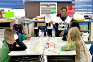 A high school student wearing a white football jersey sits in front of a group of young elementary kids and reads to them.