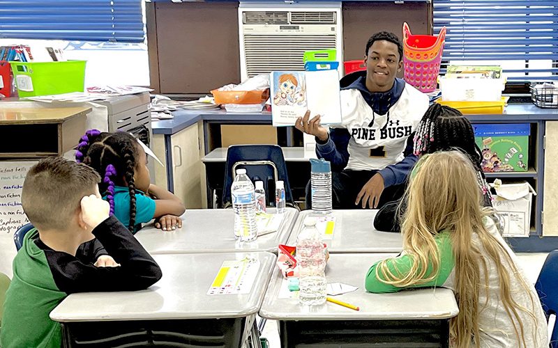 A high school student wearing a white football jersey sits in front of a group of young elementary kids and reads to them.