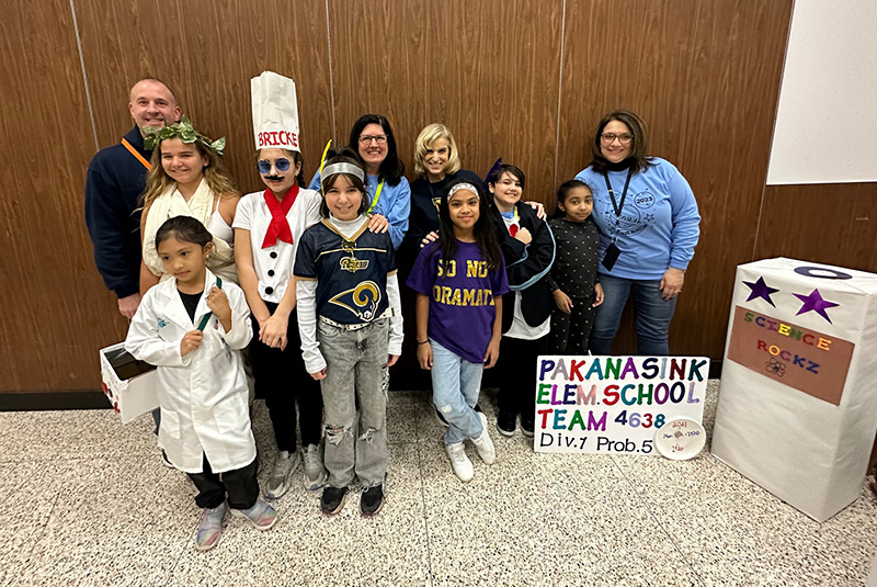 A group of seven elementary school students stand with four adults, who are all in the back. The kids are dressed in costumes for a project they are doing.