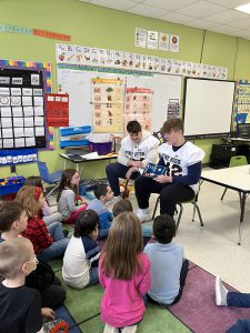 Two high school students wearing white football jerseys sit in front of a group of young elementary kids and read to them.