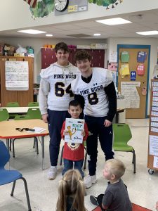 Two high school students wearing  white football jerseys stand with a little elementary student who is holding up a dr. Seuss book.