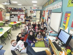 A class full of younger elementary students sit on the floor behind a high school boy lying on the floor wearing odd shaped sunglasses and a white football jersey.