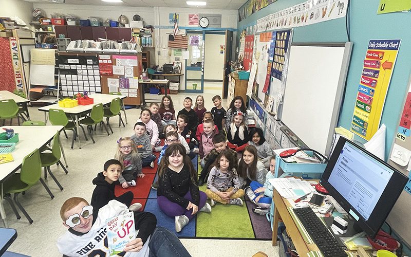 A class full of younger elementary students sit on the floor behind a high school boy lying on the floor wearing odd shaped sunglasses and a white football jersey.
