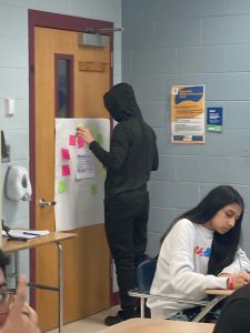 A high school girl sits at a desk writing. A high school boy stands behind her putting a post-it note on a large poster.