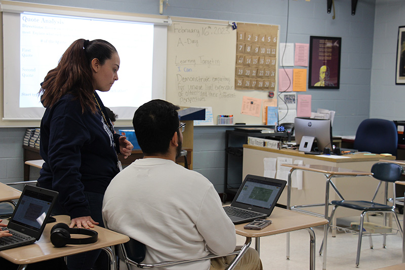 A woman with long dark hair stands next to a high school young man sitting at his desk, looking at a Chromebook.