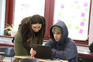 A woman with long dark hair, wearing green pants and a black shirt, leans down to a student sitting at his desk, wearing a hoodie. They are looking at a Chromebook.
