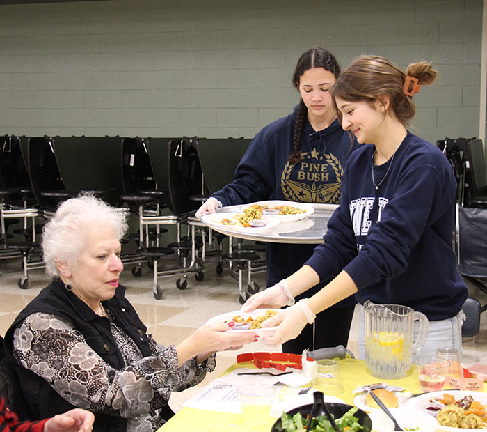 Two high school students hand a dish of food to an older woman sitting at a table.