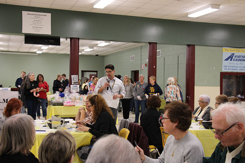 A man with dark hair and a light shirt stands in a group with a microphone and talks to the group.