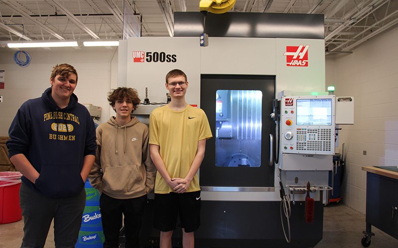 Three high school seniors stand next to a large machine.
