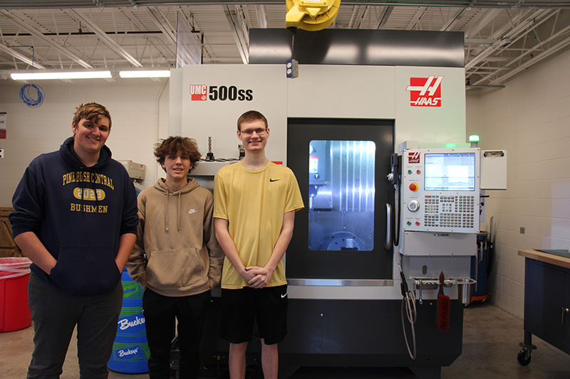 Three high school seniors stand next to a large machine.