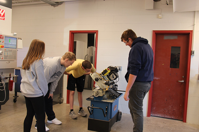 Four high school students watch while one of them works a small machine in a work shop.
