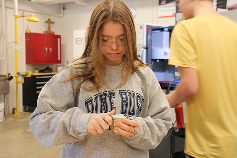 A high school senior with long blonde hair wearing a gray sweatshirt that says Pine Bush in blue, uses a sharp instrument to trim a piece of metal.