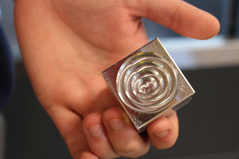 A close up of a hand holding a piece of metal that just went through a milling machine. It has cuts in it to make concentric circles.