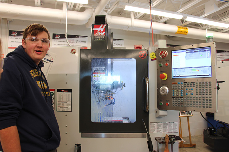A tall high school students stands next to a machine that is in the process of milling a metal.
