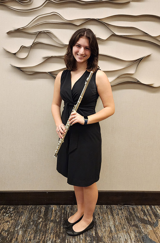 A high school senior, with long dark hair, wearing a black dress, holds her flute and smiles.