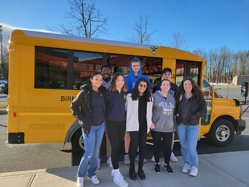 A bright blue sky in the background, eight high school students stand together and smile, with a small yellow school bus behind them.